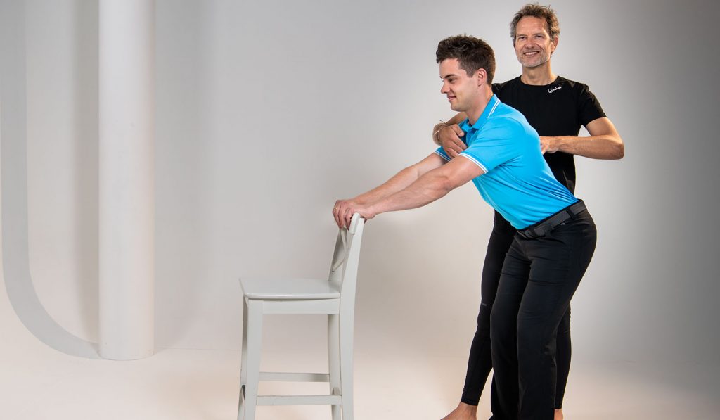 Pilatesübung: Sven Dyhr stützt sich auf Stuhllehne und Christian Lutz stabilisiert ihn