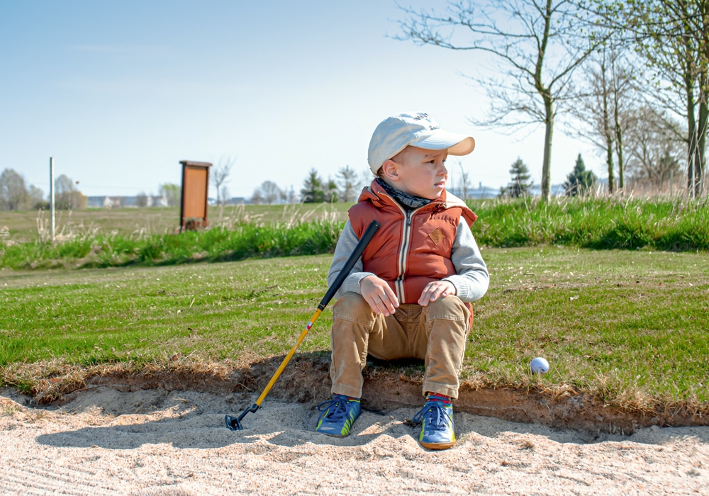 KLEINER JUNGE MIT GOLFSCHLÄGER IM SANDBUNKER